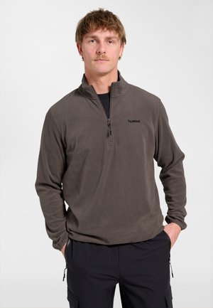 Mand med kort hår og overskæg iført en grå fleece med kvart lynlås og sorte bukser, stående med hænderne i lommerne mod en hvid baggrund.