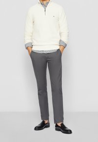 Pletený bílý svetr na polovinu s vykrojením, pod ním šedá kostkovaná košile, šedé slim-fit kalhoty a černé kožené boty. Jednoduchý, moderní a strukturovaný design.