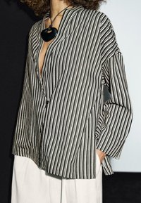 Black and white striped shirt with a deep V-neck, loose fit, and long sleeves. Textured fabric with a pocket detail on the side.