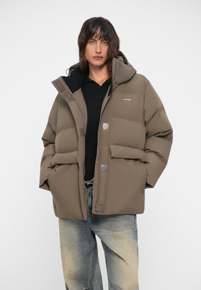 Puffer jacket in brown, featuring a hood, two front pockets, and a zipper closure with square accents. Worn over a black shirt.
