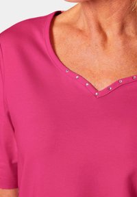 Rosa T-Shirt mit kurzen Ärmeln und V-Ausschnitt, verziert mit kleinen silbernen Nieten am Rand. Hergestellt aus glattem, atmungsaktivem Material.