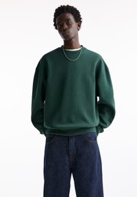 Dark green cotton sweatshirt with dropped shoulders, ribbed cuff, and hem. Paired with dark blue jeans, featuring a classic fit and minimal stitching.