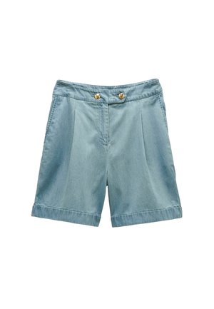 Junge Frau trägt eine gelbe Jacke über einem geblümten, in der Taille gebundenen Hemd, hellblaue Shorts und braune Slip-on-Sandalen und steht auf Teppichen.