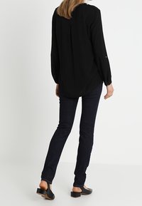 Kvinde der står med ryggen til, iført en sort langærmet bluse, mørke skinny jeans og sorte sko med lav hæl mod en hvid baggrund.