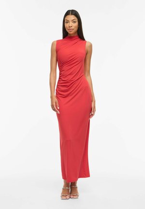 Kvinde iført ærmeløs rød ruched maxikjole med høj halsudskæring og sølvfarvede hælede sandaler, stående mod en hvid baggrund.