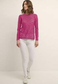 Top lungo a maniche in pizzo fuchsia con motivo floreale, scollo rotondo, abbinato a pantaloni bianchi aderenti e sneakers bianche. Design trasparente e testurizzato.