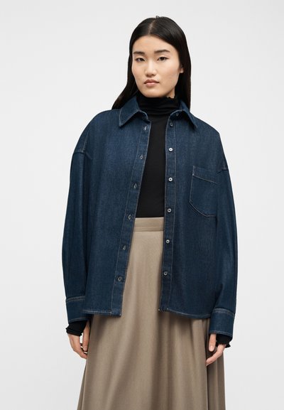 Νεαρή γυναίκα με ίσια μαύρα μαλλιά φοράει υπερμεγέθη σκούρο τζιν μπουφάν πάνω από μαύρο ζιβάγκο και μπεζ φούστα, στέκεται μπροστά σε λευκό φόντο.