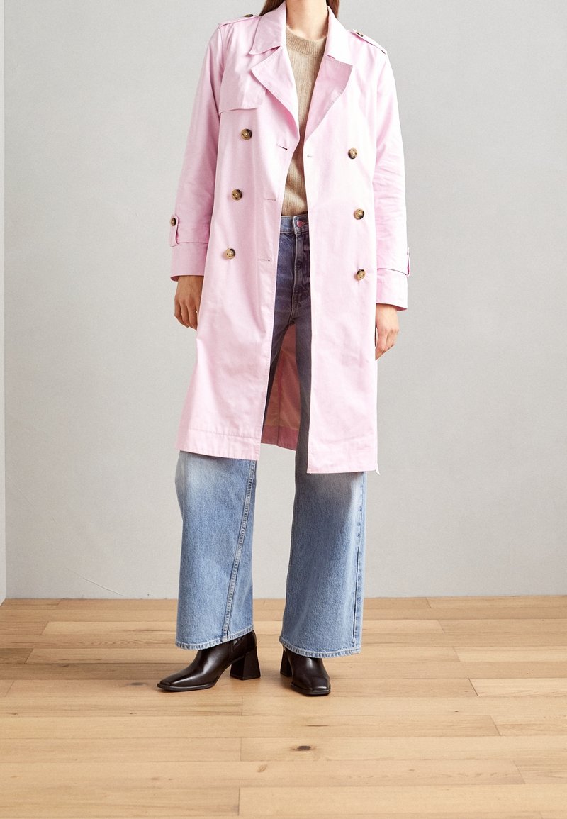 Femme portant un trench-coat à double boutonnage rose pâle sur un pull beige, jean large bleu et bottines à talons noires sur un sol en bois.