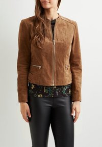 Chaqueta de ante marrón con un diseño ajustado, cierre frontal de cremallera, dos bolsillos laterales con cremallera y largo corto. Llevada sobre una blusa con estampado floral.