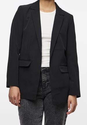 Personne portant un blazer noir sur une chemise blanche côtelée et un jean gris foncé taille haute sur fond uni.