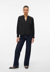 Svart blus med lång ärm och V-ringning, med samlad detalj vid kragen, kombinerad med mörkblå vida jeans och svarta platta skor.