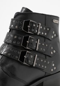 Černé kožené kotníkové boty se třemi širokými popruhy s kovovými cvočky a sponami. Hladký povrch s rovně střiženým designem a blokovým podpatkem.