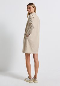 Robe chemise en lin beige avec col et manches longues, dotée de poches latérales et d'une coupe décontractée, associée à des baskets blanches.