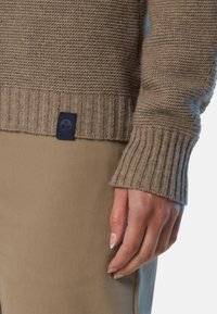 Beige gestrickter Pullover mit geripptem Saum und Bündchen, ausgestattet mit einem dunkelblauen Stoffetikett mit Logo an der Seite. Das Modell trägt beiges Hosen.