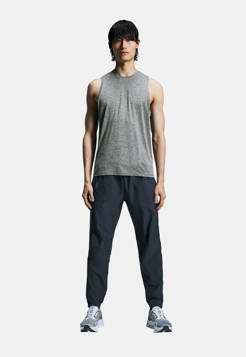 Grijze mouwloze sporttop, ruime marineblauwe broek, grijze sportschoenen met zwarte accenten, staande pose, minimalistisch ontwerp, neutrale achtergrond.
