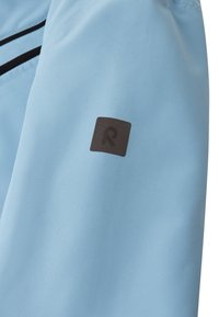 Lyseblåt stof med en glat tekstur, der har et lille sort firkantet logo med et stiliseret "R" på venstre side.
