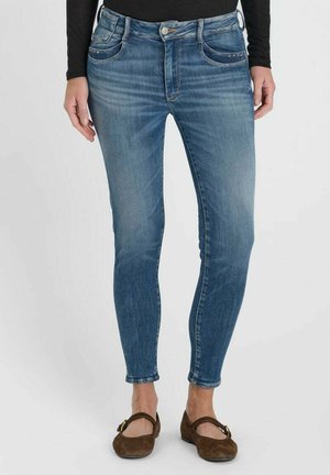 Personne portant un jean slim bleu et des chaussures plates marron, debout sur un fond clair uni.