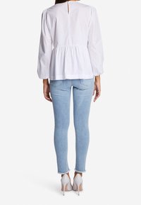 Donna che indossa una camicia bianca a maniche lunghe, jeans skinny azzurri chiari con orli sfilacciati e scarpe con tacco bianche, vista da dietro.