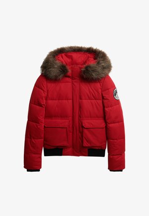 Rød puffjakke med hætte med pelskant, lynlåslukning og to forreste påsyede lommer. Har ribbede sorte manchetter og et rundt logo-patch på ærmet.