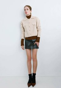 Chaqueta beige de punto con botones delanteros y dos bolsillos, combinada con un jersey de cuello alto marrón, acompañado de una minifalda negra brillante.