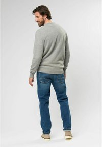 Hombre con suéter gris y jeans azules de pie, mirando por encima del hombro, frente a un fondo gris claro liso.
