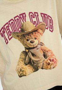 Beige t-shirt met een afbeelding van een teddybeer die een cowboyhoed en sjaal draagt, met de tekst "TEDDY CLUB" in kastanjebruin erboven.