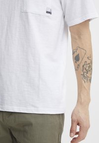 Forearm tattoo showing playing cards and a smoking woman, worn by a person in a white t-shirt and olive green pants.