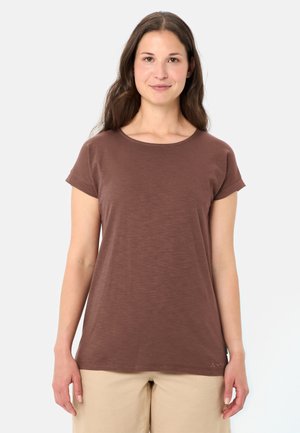 Donna con capelli lunghi castani che indossa una maglietta marrone a maniche corte e pantaloni beige, in piedi contro uno sfondo chiaro uniforme.