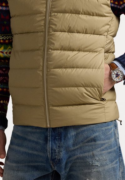 Beige puffer vest with horizontal quilted sections, front zipper, and side pockets. Worn over a patterned sweater, paired with blue jeans.