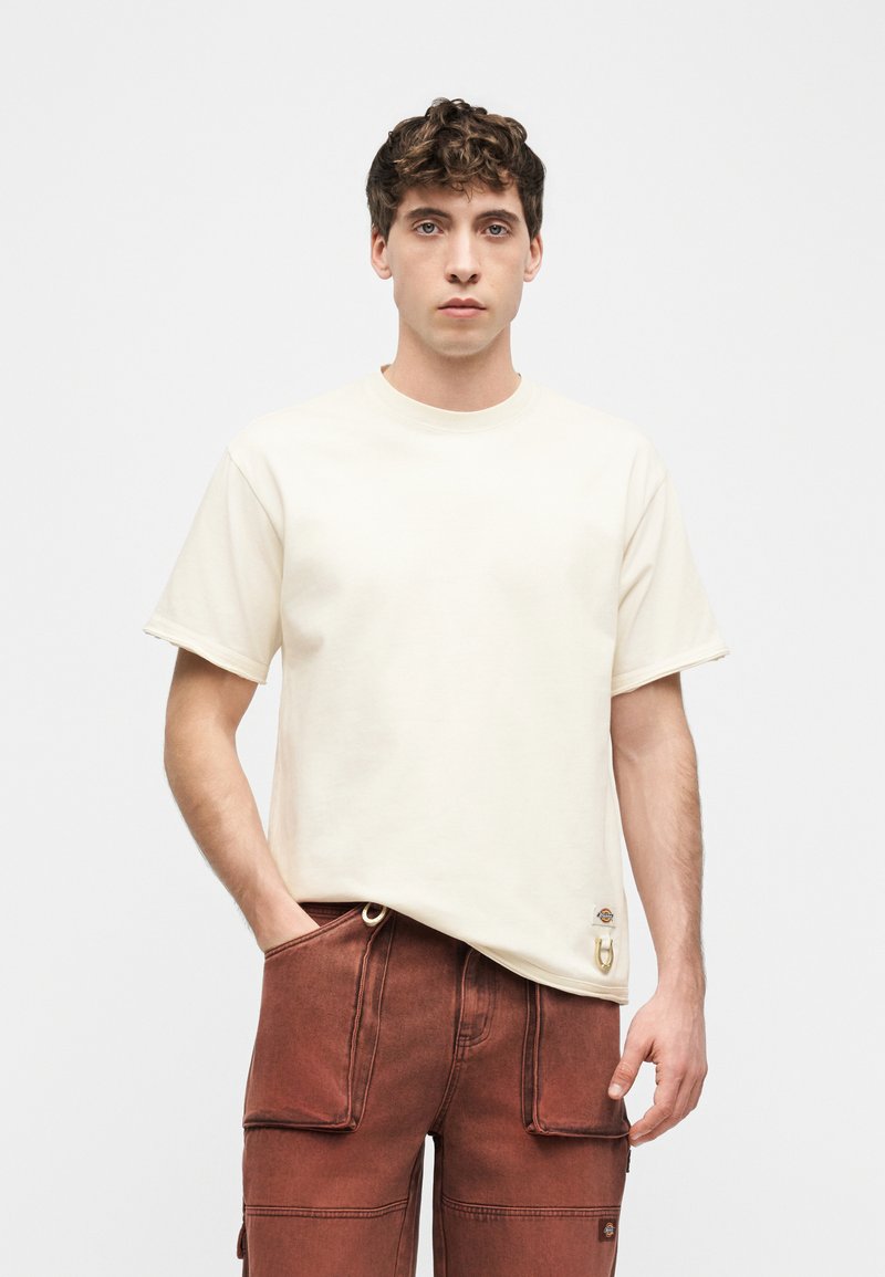 Junger Mann mit lockigem Haar, der ein schlichtes, cremefarbenes T-Shirt und rostfarbene Cargohosen trägt und vor einem weißen Hintergrund steht.