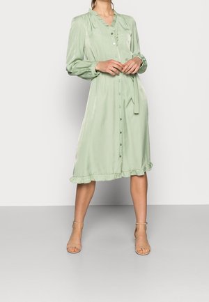 Kobieta ubrana w jasnorozzieloną, długorękawową sukienkę zapinaną na guziki, z falbankami przy kołnierzu i u dołu, stojąca w beżowych szpilkach na kostkę z paskiem.