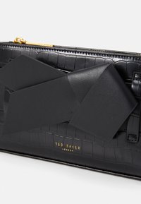 Bolsa de material preto com relevo de crocodilo, apresentando um laço preto proeminente. Logotipo dourado "Ted Baker London" na parte inferior. Fecho com zíper.