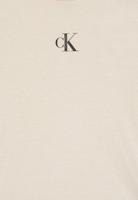 Világos bézs pamut póló, a bal felső részen egy kis fekete "cK" logóval. Az anyag sima textúrájú.