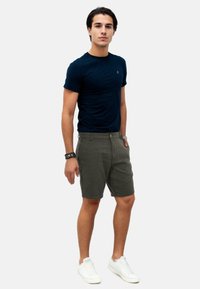 Junger Mann steht mit einer Hand in der Tasche, trägt ein navyblaues T-Shirt, olivgrüne Shorts, weiße Sneakers und schwarze Armbänder vor einem weißen Hintergrund.