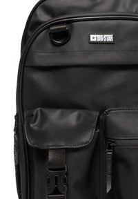 Mochila negra hecha de un material suave, con múltiples bolsillos, un cierre de hebilla y un parche de logo cosido en la parte frontal.