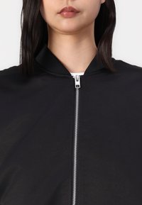 Fekete bomber dzseki bordázott gallérral és ezüst cipzárral. Az anyag simának tűnik, minimalista dizájnnal és látható minták nélkül.