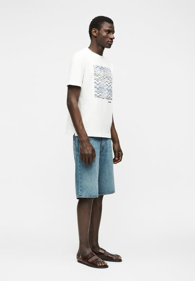 Άνδρας όρθιος, φορεί λευκό μπλουζάκι με γραφικά, μπλε τζιν σορτς και καφέ σανδάλια, με λευκό φόντο.