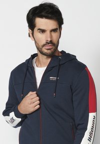 Jūros pajėgų zip-up džemperis su raudonais ir baltais akcentais, su raišteliu reguliuojama kapuce, logotipu ant krūtinės, gumuotomis rankovėmis ir priekinėmis užtrauktukinėmis kišenėmis.