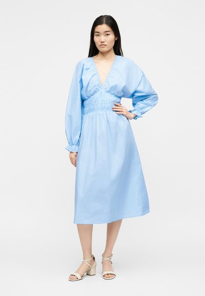 Γυναίκα με μακριά σκούρα μαλλιά φοράει ανοιχτό μπλε midi φόρεμα με μακριά μανίκια και βαθύ ντεκολτέ σε σχήμα V, καθώς και εκρού σανδάλια με τακούνι, στέκεται με το χέρι στη μέση.