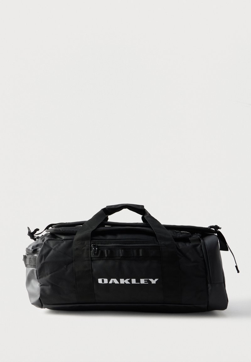 Μαύρη τσάντα Oakley duffel με επάνω λαβές, εμπρόσθια τσέπη με φερμουάρ και λογότυπο σε απλό φωτεινό φόντο.