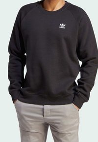 Osoba v černém svršku Adidas s logem na hrudi a světle šedých kalhotách, s rukama v kapsách.