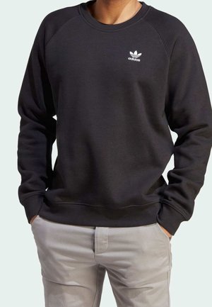 Osoba v černém svršku Adidas s logem na hrudi a světle šedých kalhotách, s rukama v kapsách.