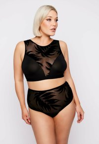 Eine kurvenreiche Frau trägt schwarze, durchsichtige Lingerie mit Blattmustern und blickt zur Seite vor einem schlichten, hellen Hintergrund.