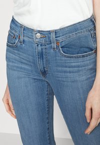 Jeans din denim de culoare albastru deschis, cu un design mulat, cu fermoar și închidere cu buton, având cinci buzunare și o decolorare subtilă în material.