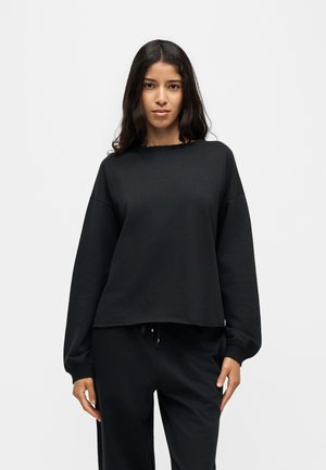 Femme aux longs cheveux foncés portant un haut noir ample à manches longues et un pantalon noir, debout devant un fond clair uni.