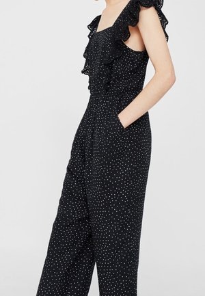 Ærmeløs sort jumpsuit med hvide prikker, flæsekrave og sidelommer båret af en person, der står mod en lys baggrund.