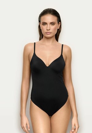 Mujer con el pelo liso y mojado, que lleva un traje de baño negro de una pieza ajustado con tirantes finos sobre un fondo claro y simple.