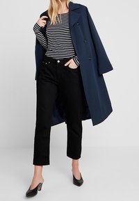 Trench coat blu navy su una maglietta a maniche lunghe a righe bianche e nere, abbinato a pantaloni neri a gamba dritta e tacchi kitten neri.