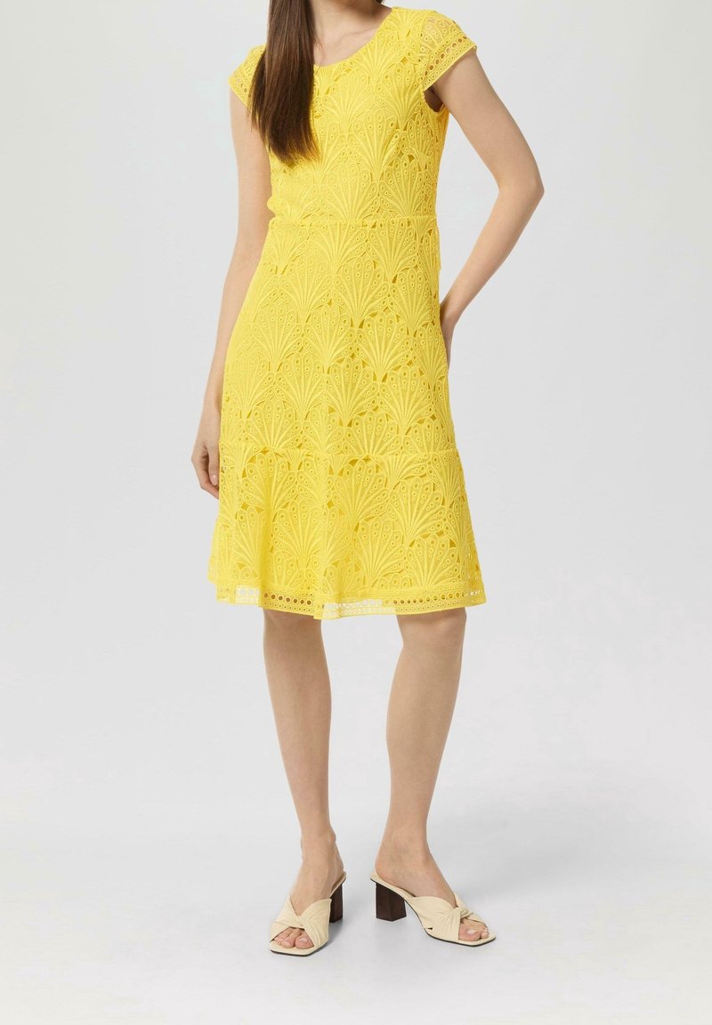 Femme portant une robe en dentelle jaune longueur genoux avec manches courtes et des sandales beiges à talons, debout devant un fond uni.