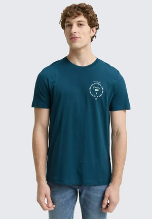Camiseta de algodón en color azul verdoso con mangas cortas, cuello redondo y un diseño circular impreso en el pecho en color blanco, llevada con jeans de color azul claro.